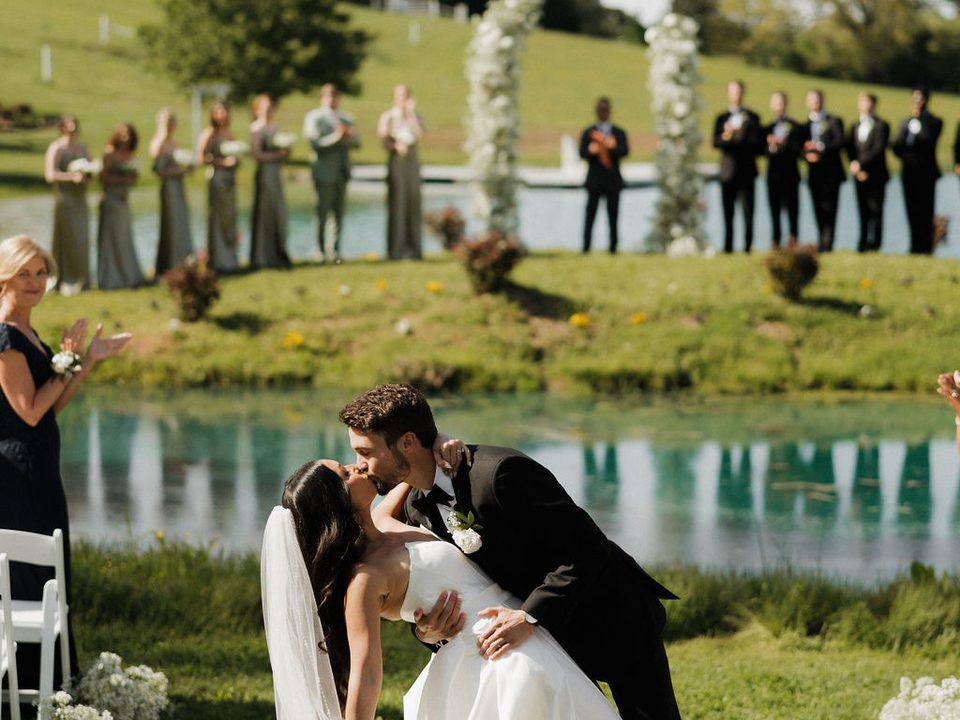 A groom dips his bride mid-kiss on the pond island, the wedding party lined along the water at Cedar Pond Farms near Sevierville