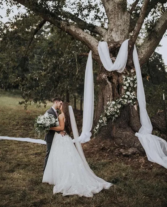 Una novia bajo el Gran Roble adornado con tela blanca y flores en una boda en granja cerca de Gatlinburg