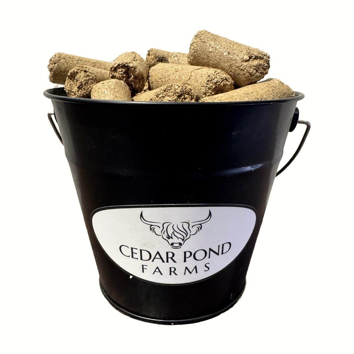 Black metal bucket labeled Cedar Pond Farms filled with compressed feed logs against a white background.
