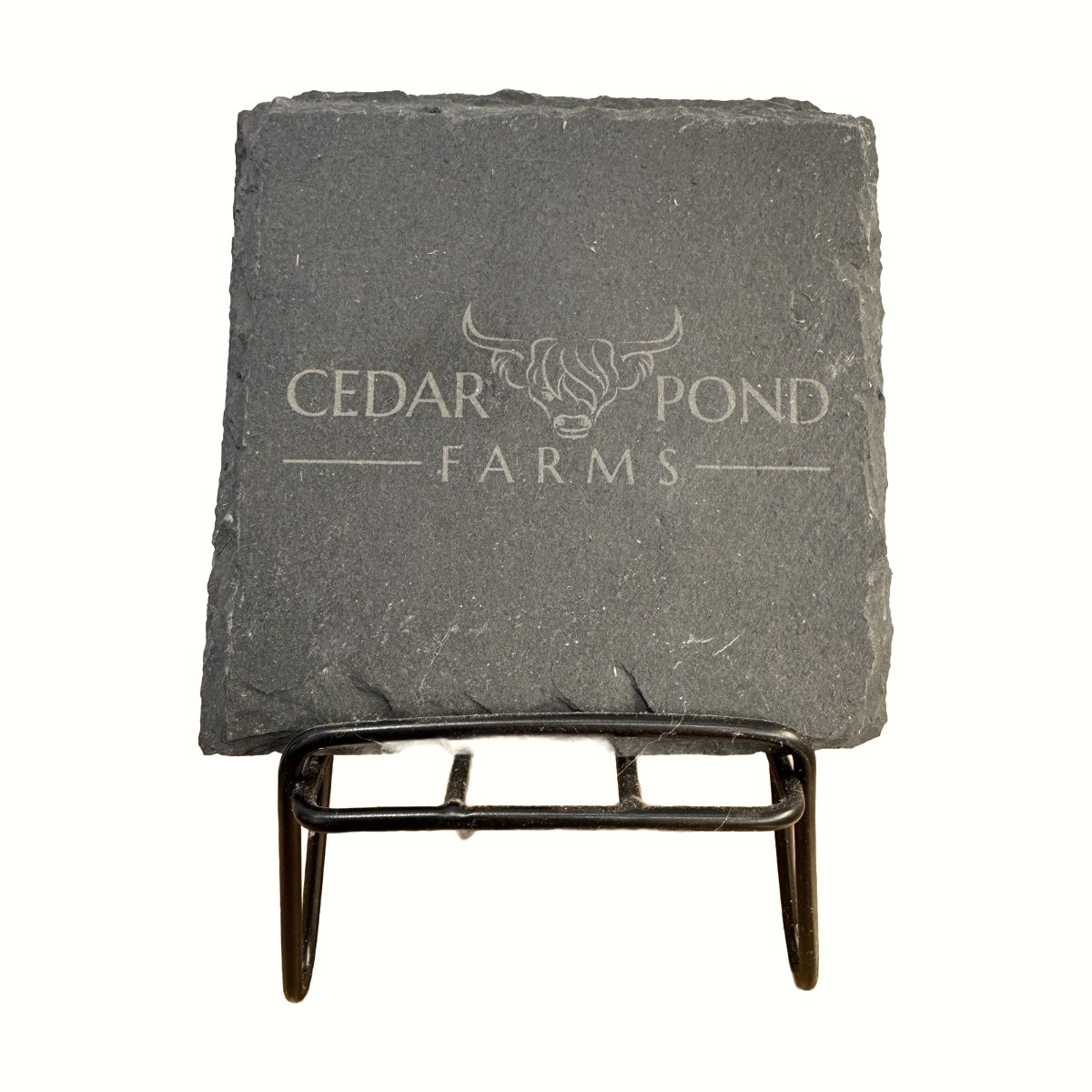Square gray slate coaster engraved with the Cedar Pond Farms logo featuring a highland cow, displayed on a black metal stand.