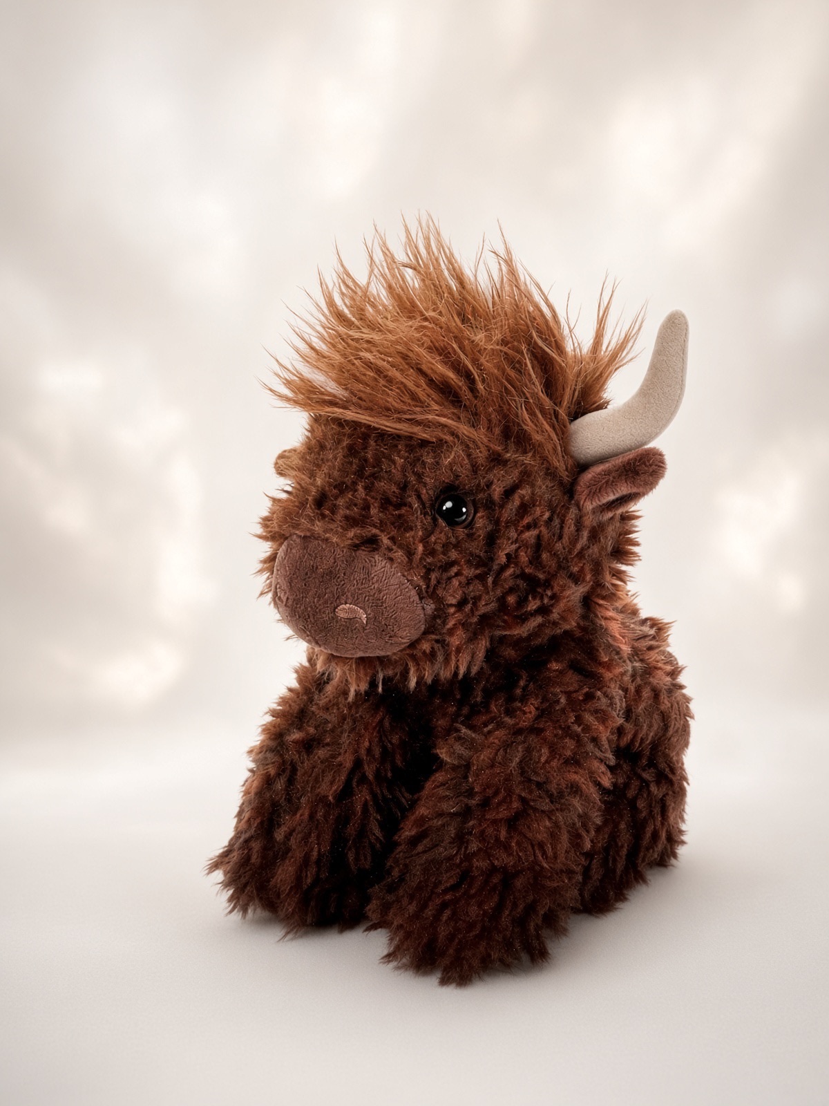 A 9 inch plush Highland cow toy with shaggy chocolate-brown fur, small horns, and a fuzzy fringe, sitting against a soft backdrop.