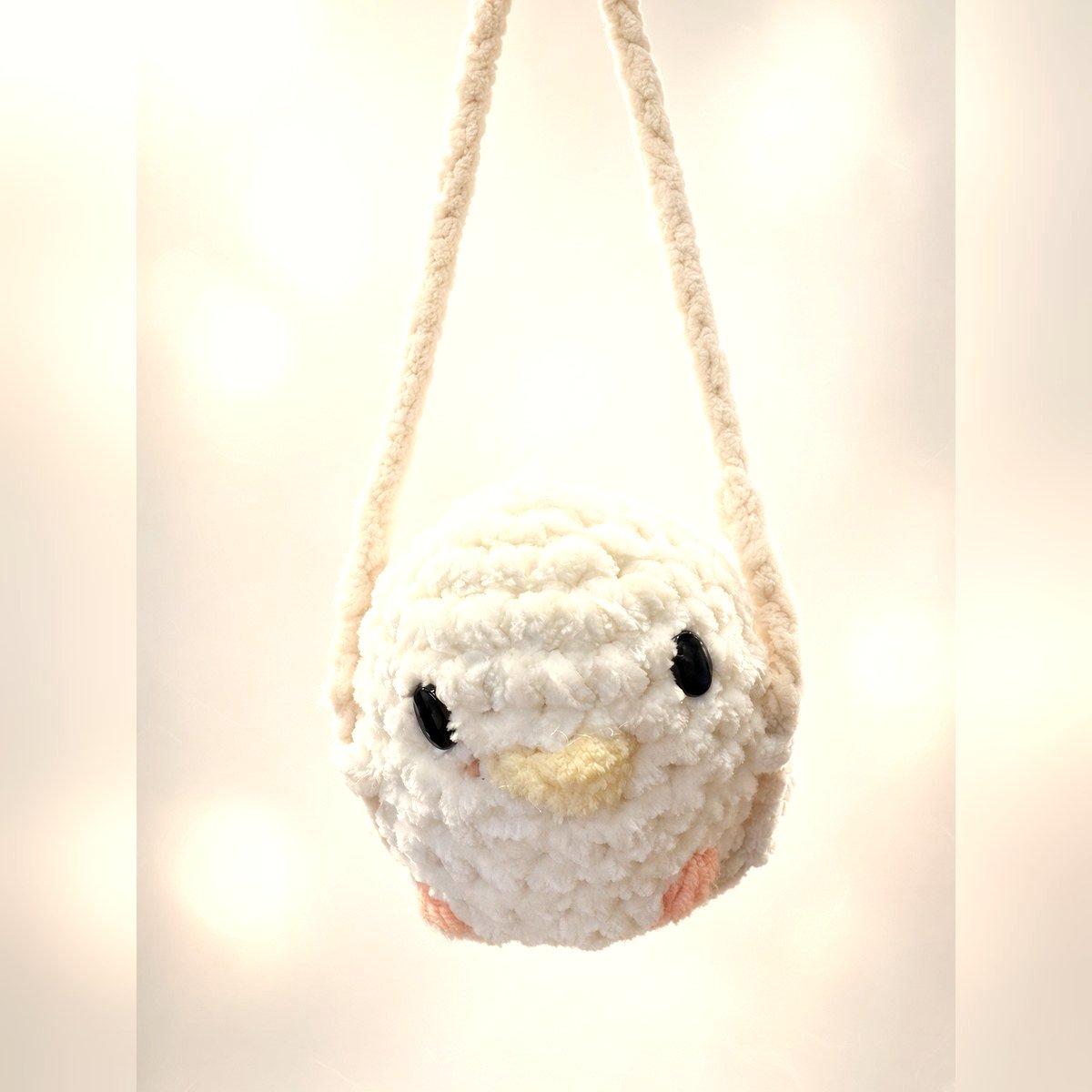 A small round crocheted chick-shaped bag in white yarn with a yellow beak and black eyes, hanging from a matching crocheted strap.