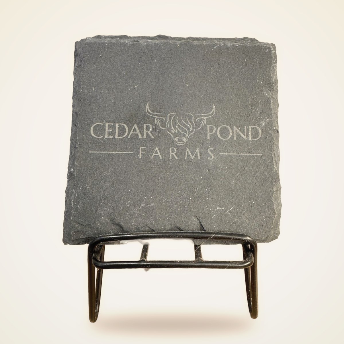 Square gray slate coaster engraved with the Cedar Pond Farms logo featuring a highland cow, displayed on a black metal stand.